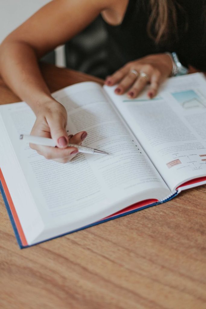 Close-up of an adult woman studying with a pen in hand, reading a textbook at a wooden table.